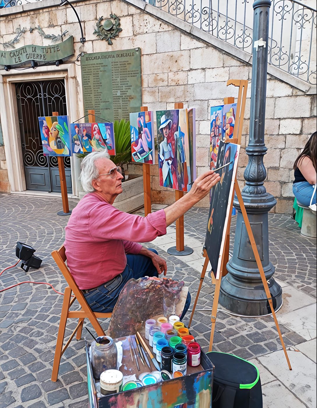 Giacomo De Troia dipinge al cavalletto in piazza con i dipinti esposti alle spalle